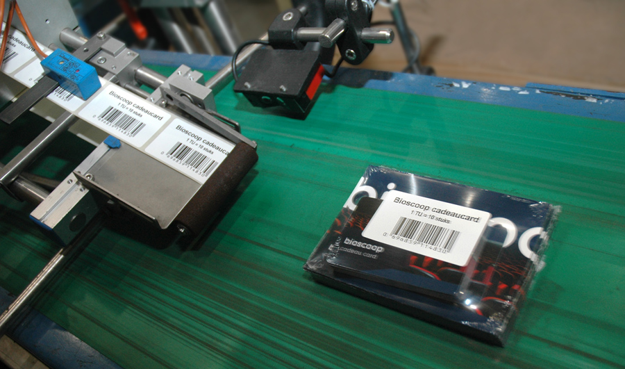 A conveyor belt with a black package labeled "Bioscoop cadeaukaart." Nearby, a machine applies barcode labels, suggesting an industrial setting.