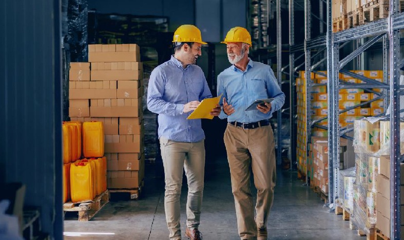 Warehouse workers reviewing inventory and logistics data in a distribution center aisle with storage racks and packaged goods.