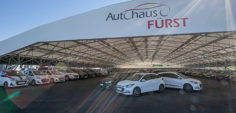 A large car dealership canopy displaying a variety of cars parked underneath, including two prominent white cars in front. Sunlight creates lens flares.