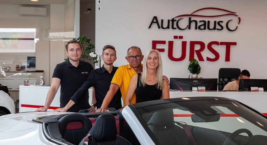 A smiling group stands in a car showroom in front of a white convertible. The backdrop features "Autohaus Fürst" signage. The ambiance is welcoming.