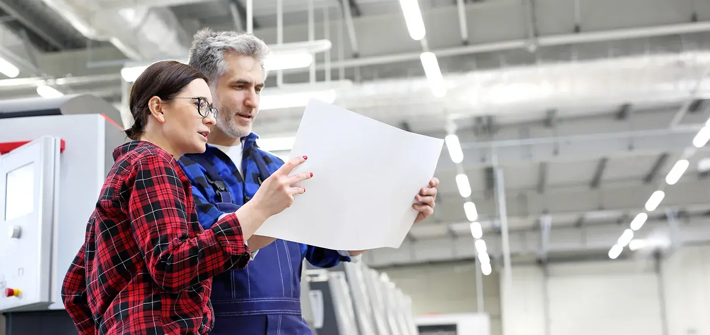  A male and a female industrial worker are standing in a large production facility, looking at a blank sheet of paper they are holding between them. The woman is wearing glasses and a red and black plaid shirt, while the man is in a blue work suit. Heavy machinery is visible in the blurred background.