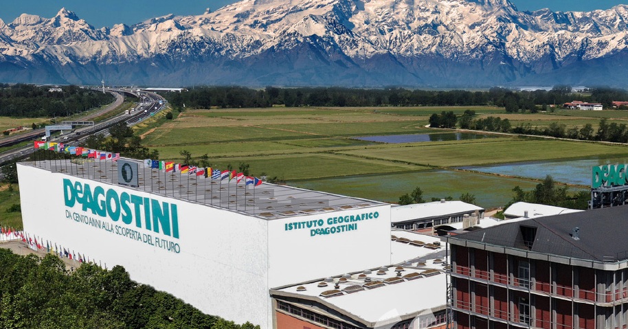 Aerial view of DeAgostini Publishing headquarters in Italy, showing the Istituto Geografico DeAgostini building with surrounding landscape and Alpine backdrop.