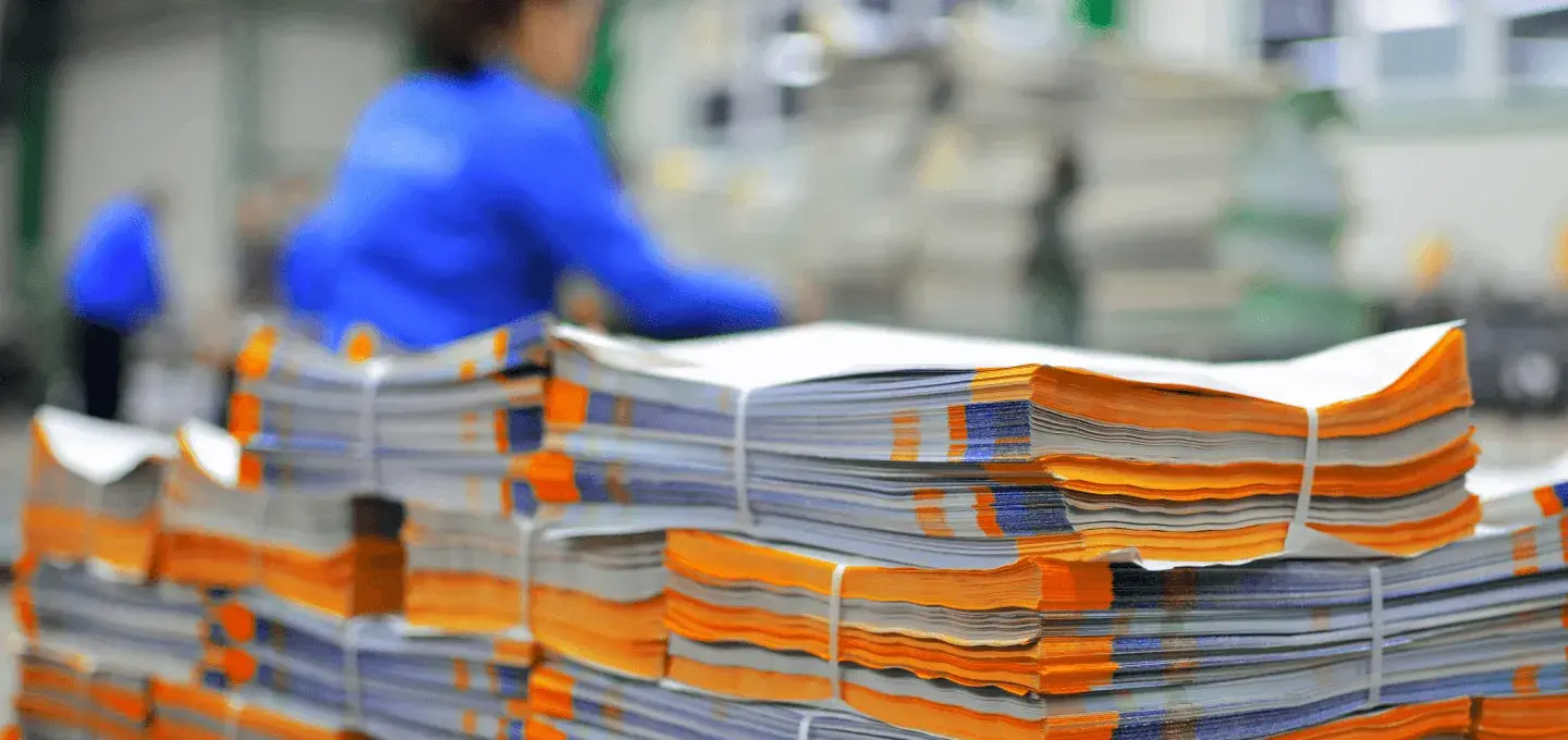 Stacks of folded paper with orange and blue accents are tied together in a print shop. A worker in a blue uniform is blurred in the background.