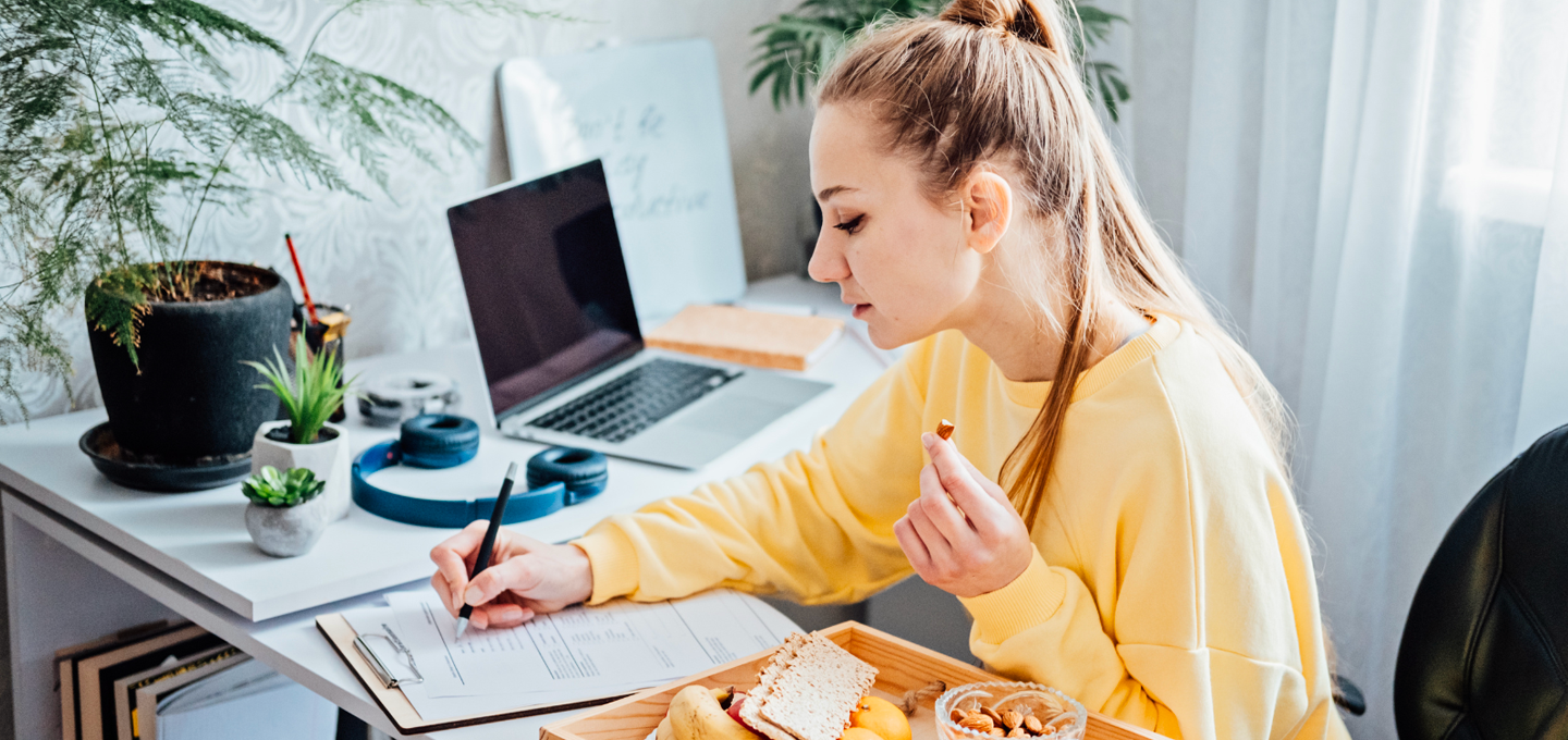A woman in a yellow sweater sits at a desk, writing on a clipboard while eating. A laptop and plants are on the desk, conveying a focused, homey atmosphere.
