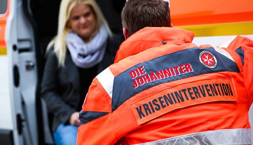 Emergency worker in orange Johanniter crisis response jacket speaking to person beside ambulance
