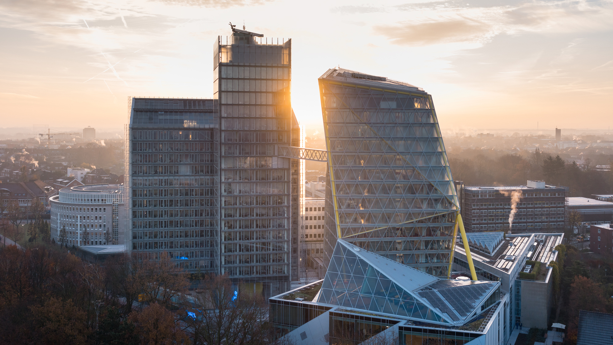 Modern office towers connected by a skybridge at sunrise, featuring contemporary glass architecture and sustainable design, set against a misty urban skyline in soft morning light.