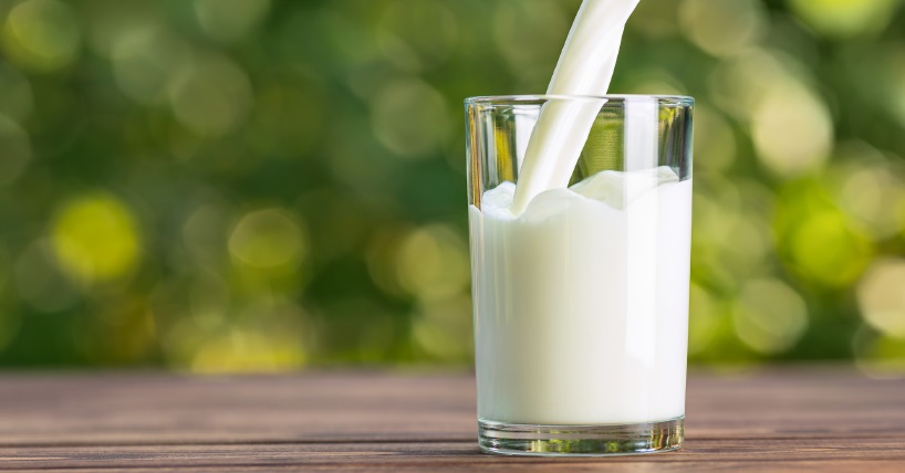 A stream of white milk is being poured into a clear glass sitting on a wooden surface. The background is softly blurred with green and yellow hues, suggesting an outdoor or natural setting.