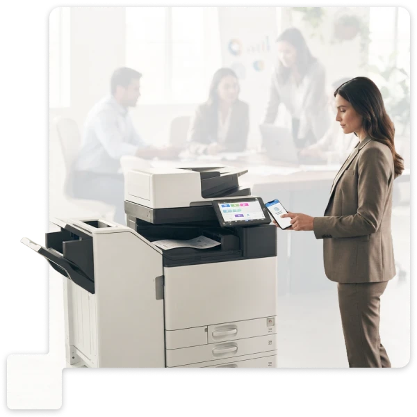 Office worker using a multifunction printer, with colleagues meeting around a table in the background.