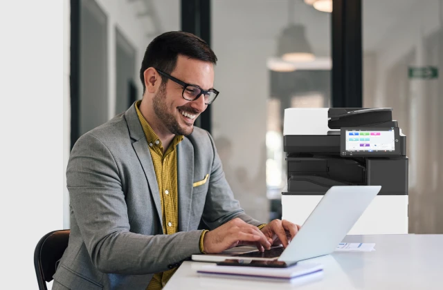  A man in glasses and a grey suit jacket sits at a white table, smiling as he types on a laptop. To his left, a Ricoh printer is visible. The background is a bright office space.