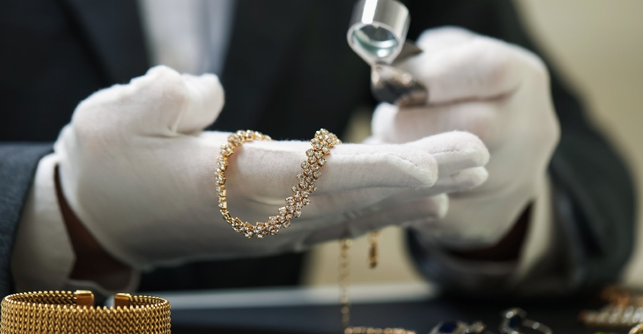 A jeweler wearing white gloves examines a diamond bracelet with a loupe. The scene conveys precision and luxury, with a focus on the meticulous inspection.