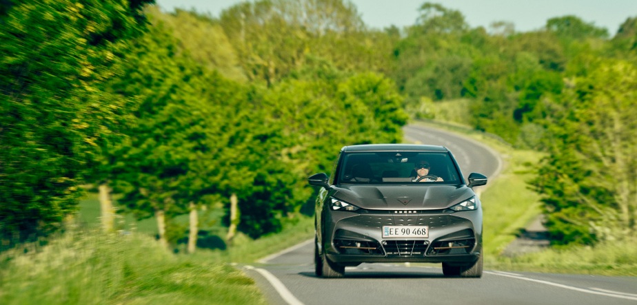 Photograph of a black electric car driving on a winding rural road surrounded by green trees and grass. The car is centered in the frame, with a clear view of its front and license plate, highlighting its sleek design and modern headlights.