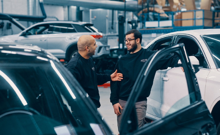 Photograph of two men engaged in conversation inside a car dealership or showroom, surrounded by several vehicles. The setting features modern cars with one man gesturing while the other listens, highlighting a friendly and professional interaction.