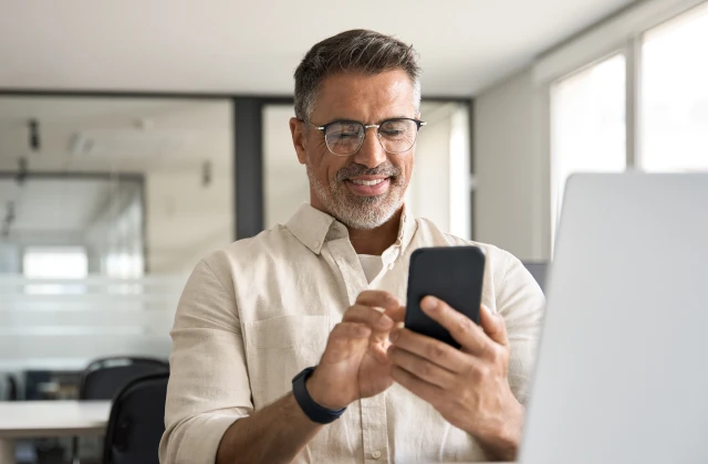 A smiling man in glasses and a beige shirt is sitting at a desk in a modern office, using his smartphone while working on a laptop. A multifunction printer with a touchscreen interface is positioned nearby. The office has large windows and glass partitions, creating a bright and professional atmosphere.
