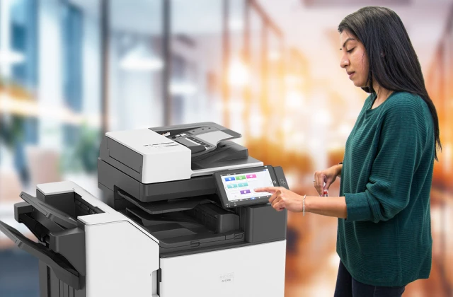 A woman in a green sweater is using a modern multifunction printer with a touchscreen interface in a well-lit office environment. She is interacting with the device, pressing options on the screen while holding a card in her other hand. The background is blurred, creating a professional and focused setting.