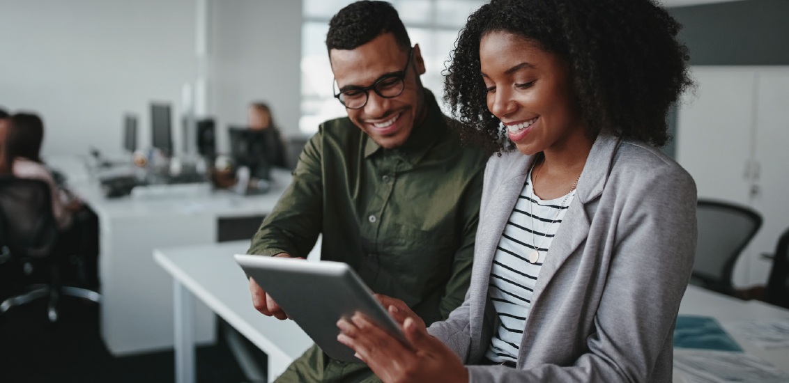 Two colleagues in an office smile while looking at a tablet. The workspace is modern, with computers in the background, creating a collaborative atmosphere.