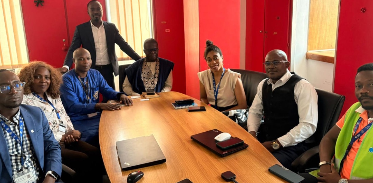 A diverse group of eight individuals sits around a wooden conference table in a meeting room with red walls, conveying a professional and collaborative atmosphere.