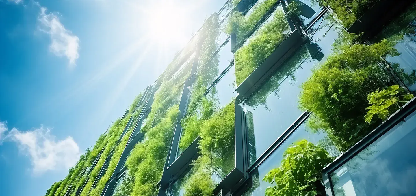 A large, modern building with a glass facade is covered in lush green plants, which hang from and grow on the exterior. The sun shines brightly from above, creating a lens flare and illuminating the building against a blue sky with scattered clouds.