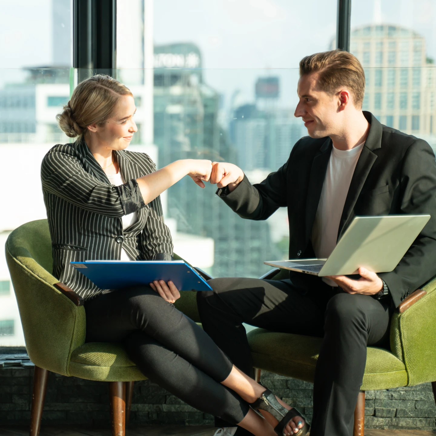 Two professionals seated in an office, exchanging a fist bump while holding a laptop and folder.