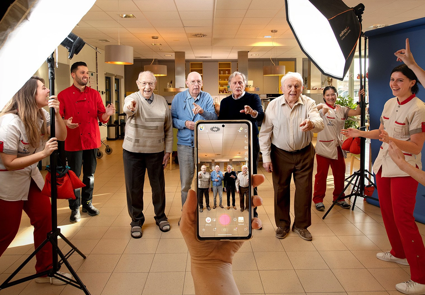 A group of elderly people and caregivers smile and pose for a photo in a well-lit room with professional lighting. A hand holds up a phone capturing the scene.